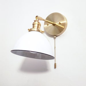 May include: A white and gold wall sconce with a pull chain switch. The sconce has a white, dome-shaped shade and a gold metal base.