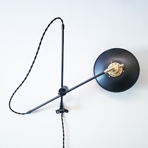May include: Black wall-mounted lamp with a gold-colored socket and a black shade. The lamp has a long, adjustable arm and a black cord.
