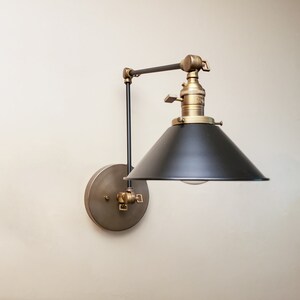 May include: A black and brass wall-mounted light fixture with a black metal shade and a brass arm. The light is adjustable and can be moved up and down.