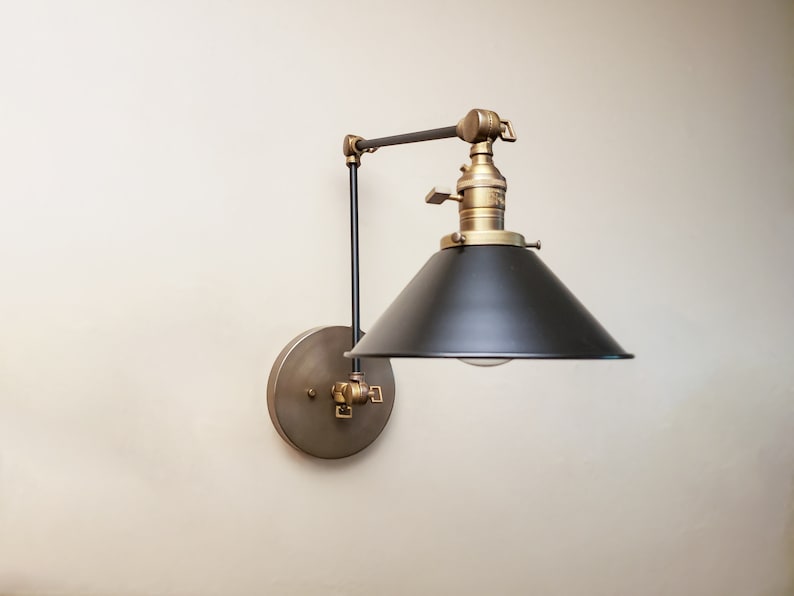 May include: A black and brass wall-mounted light fixture with a black metal shade and a brass arm. The light is adjustable and can be moved up and down.