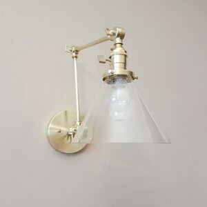 May include: A gold-toned wall sconce with a clear glass cone shade. The sconce has an adjustable arm and a round base.