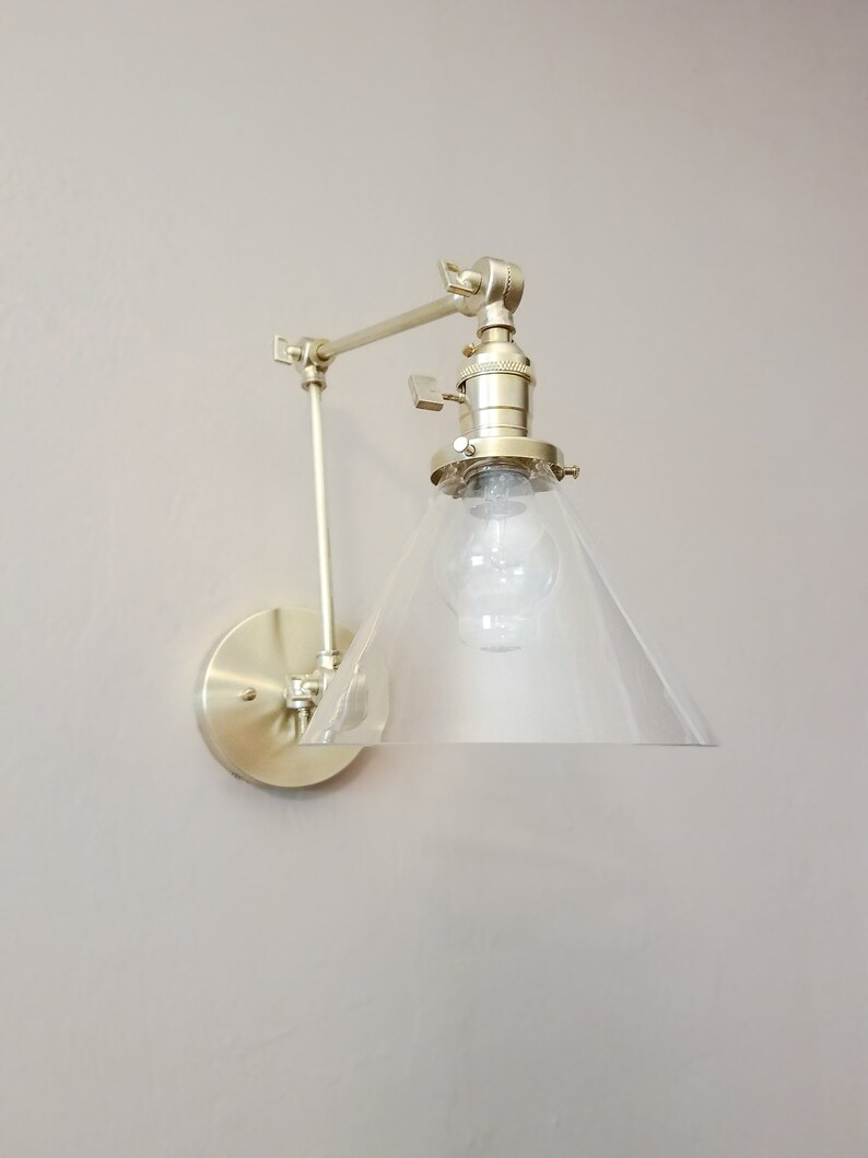 May include: A gold-toned wall sconce with a clear glass cone shade. The sconce has an adjustable arm and a round base.