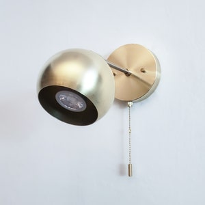 May include: A gold-toned wall sconce with a round, spherical shade and a pull chain. The sconce is mounted on a white wall.