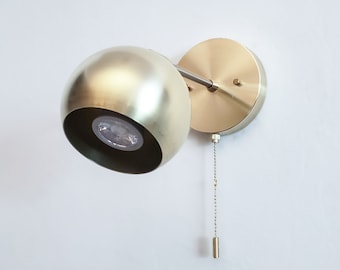 May include: A gold-toned wall sconce with a round, spherical shade and a pull chain. The sconce is mounted on a white wall.