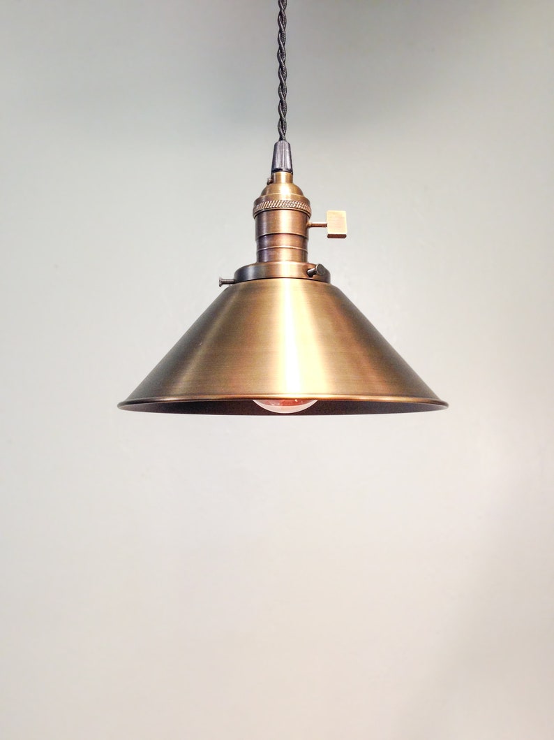Puede incluir: Una l&aacute;mpara colgante de lat&oacute;n con una pantalla en forma de cono. La luz cuelga de un cable negro.