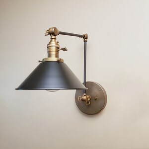 May include: A black and brass wall-mounted light fixture with a black metal shade and a swivel arm. The light fixture has a vintage industrial style.