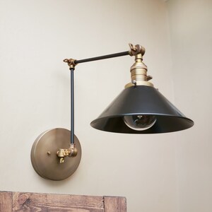 May include: A black and brass wall-mounted light fixture with a black cone-shaped shade and a single light bulb. The fixture has a swivel arm that allows the light to be directed in different directions.