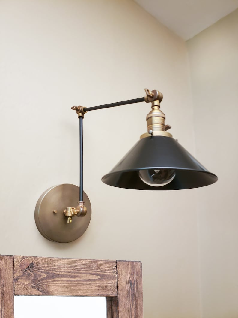May include: A black and brass wall-mounted light fixture with a black cone-shaped shade and a single light bulb. The fixture has a swivel arm that allows the light to be directed in different directions.