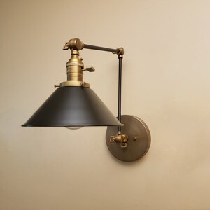 May include: A black and brass wall-mounted light fixture with a black metal shade and a brass arm. The light fixture is adjustable and can be moved up and down.