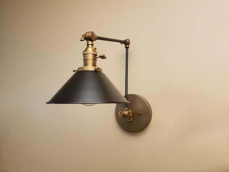 May include: A black and brass wall-mounted light fixture with a black metal shade and a brass arm. The light fixture is adjustable and can be moved up and down.