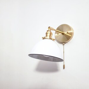 May include: A white and gold wall sconce with a round shade and a pull chain. The sconce is mounted on a white wall.