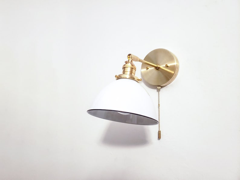May include: A white and gold wall sconce with a round shade and a pull chain. The sconce is mounted on a white wall.