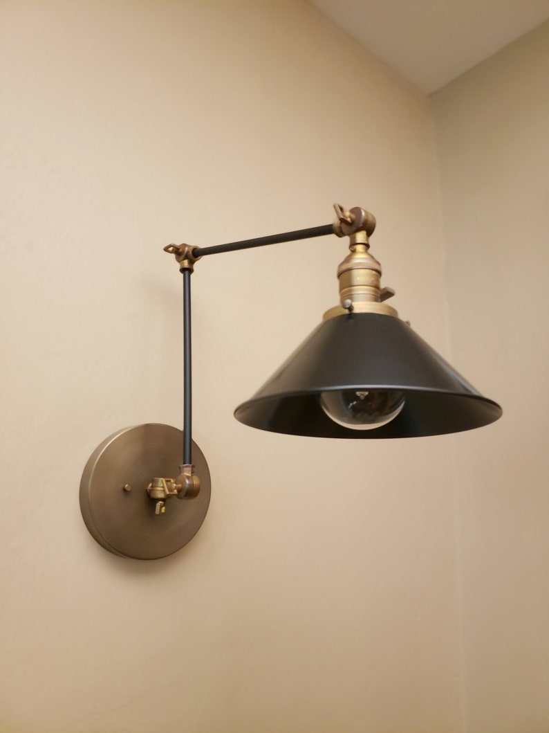 May include: A black and brass wall-mounted light fixture with a black shade and a round brass base. The light is on and the bulb is visible.