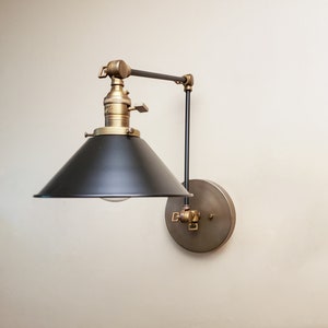 May include: A black and brass wall-mounted light fixture with a black metal shade and a brass arm. The light fixture is adjustable and can be moved up and down.