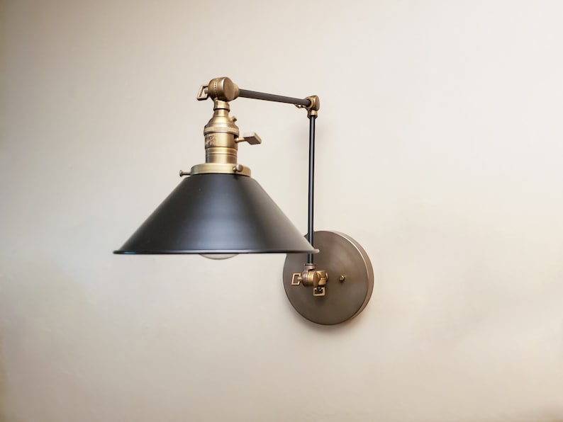 May include: A black and brass wall-mounted light fixture with a black metal shade and a brass arm. The light fixture is adjustable and can be moved up and down.