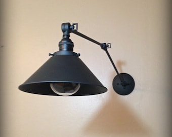 May include: Black wall-mounted light fixture with a black metal shade and a single light bulb. The fixture has a long, adjustable arm.