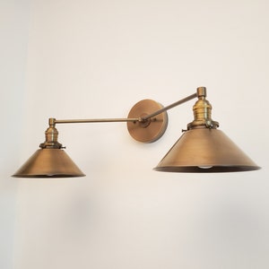 May include: A brass-toned, two-light wall sconce with adjustable arms and conical shades. The fixture is mounted on a circular backplate and features a vintage-inspired design, suitable for interior lighting.
