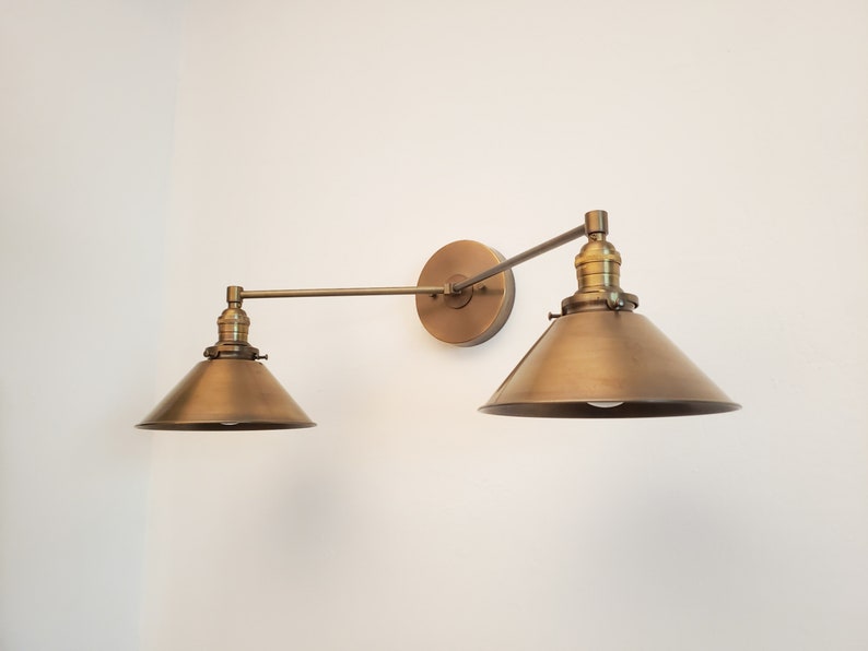 May include: A brass-toned, two-light wall sconce with adjustable arms and conical shades. The fixture is mounted on a circular backplate and features a vintage-inspired design, suitable for interior lighting.
