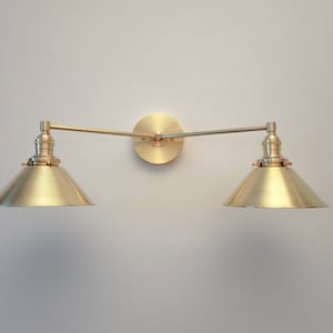 May include: A brass-toned wall-mounted light fixture with two conical shades. The fixture has a circular base and adjustable arms. The shades are angled downward, and the fixture is mounted on a light-colored wall.
