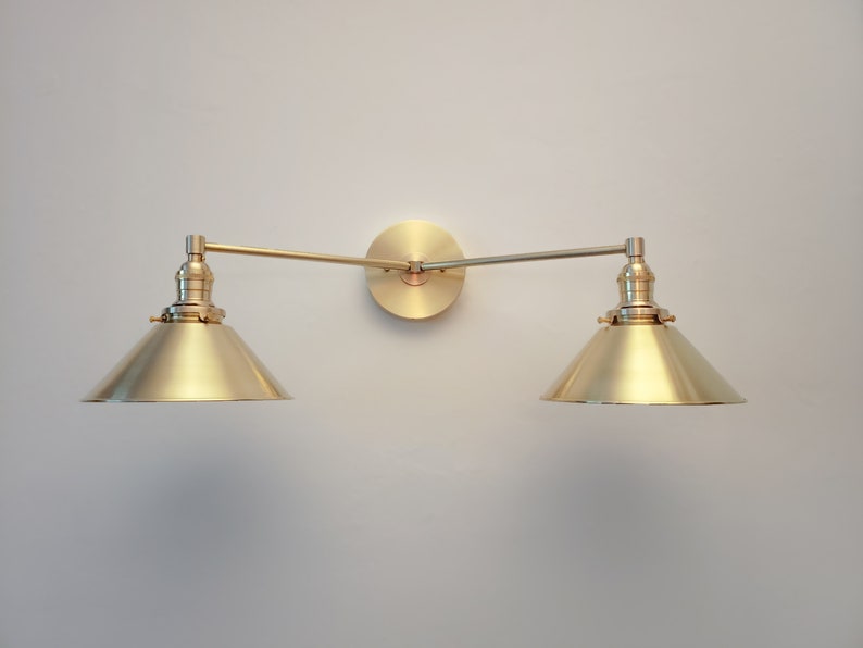 May include: A brass-toned wall-mounted light fixture with two conical shades. The fixture has a circular base and adjustable arms. The shades are angled downward, and the fixture is mounted on a light-colored wall.