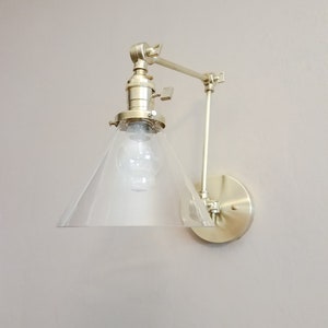 May include: A gold-toned wall sconce with a clear glass cone shade and an adjustable arm. The sconce has a simple, modern design and would be a great addition to any home.