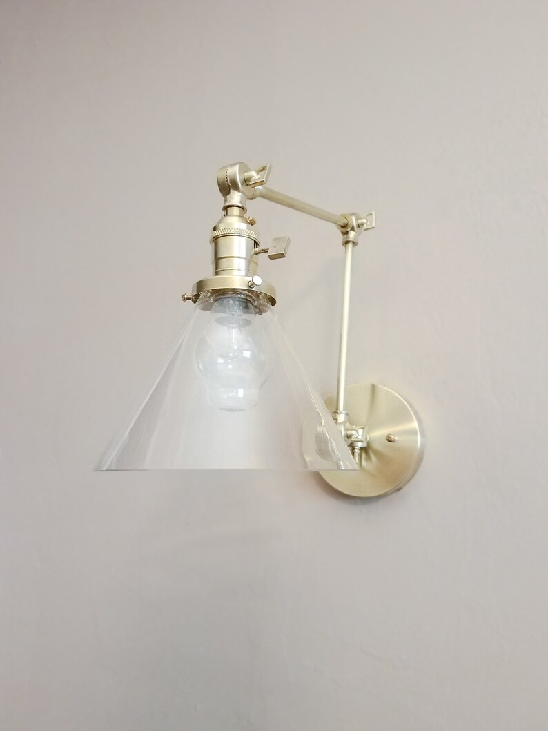 May include: A gold-toned wall sconce with a clear glass cone shade and an adjustable arm. The sconce has a simple, modern design and would be a great addition to any home.