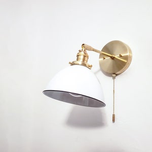 May include: A white and brass wall-mounted light fixture with a pull chain switch. The light fixture has a white, dome-shaped shade and a brass arm that extends from the wall.