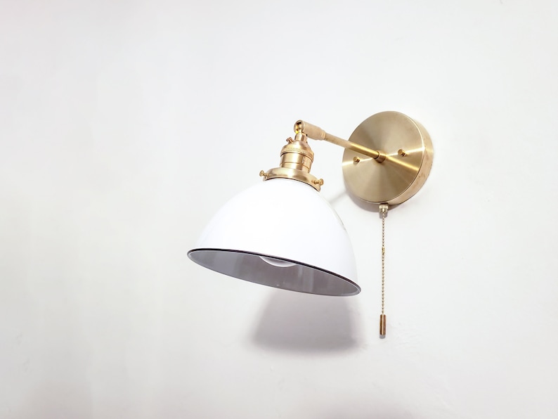 May include: A white and brass wall-mounted light fixture with a pull chain switch. The light fixture has a white, dome-shaped shade and a brass arm that extends from the wall.