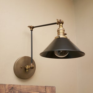 May include: A brass and black wall-mounted light fixture with a swivel arm and a black cone-shaped shade. The light fixture is mounted on a beige wall.