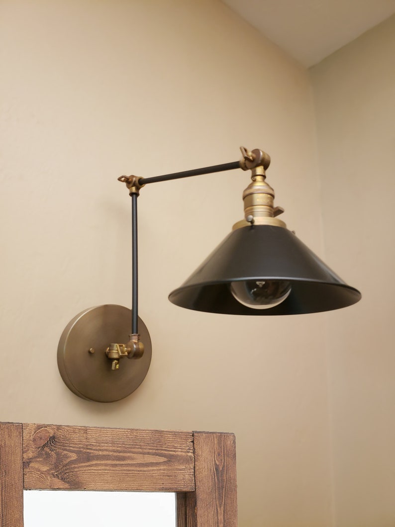 May include: A brass and black wall-mounted light fixture with a swivel arm and a black cone-shaped shade. The light fixture is mounted on a beige wall.
