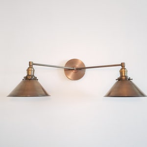 May include: A bronze-toned, two-light wall sconce with a circular backplate and adjustable arms. Each light features a conical shade. The fixture is mounted on a white wall, showcasing a vintage-inspired design.