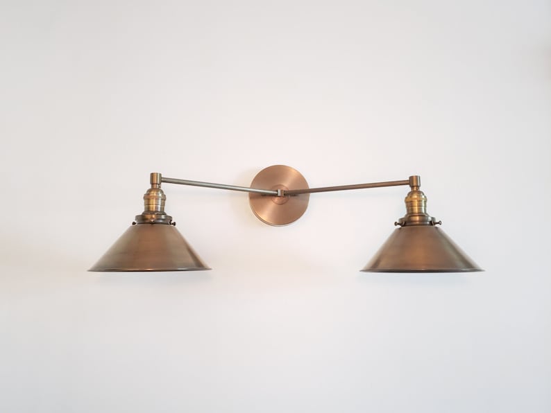 May include: A bronze-toned, two-light wall sconce with a circular backplate and adjustable arms. Each light features a conical shade. The fixture is mounted on a white wall, showcasing a vintage-inspired design.