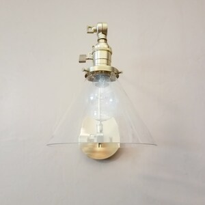 May include: A gold-toned wall sconce with a clear glass cone shade. The sconce has a simple design and is perfect for adding a touch of modern style to any room.