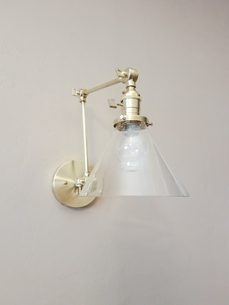 May include: A gold-toned wall sconce with a clear glass cone shade. The sconce has a swivel arm and a round base.