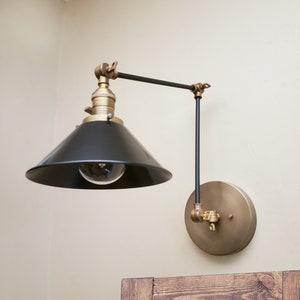 May include: A black and brass wall-mounted light fixture with a black cone-shaped shade and a round brass base. The light is adjustable and can be moved up and down.