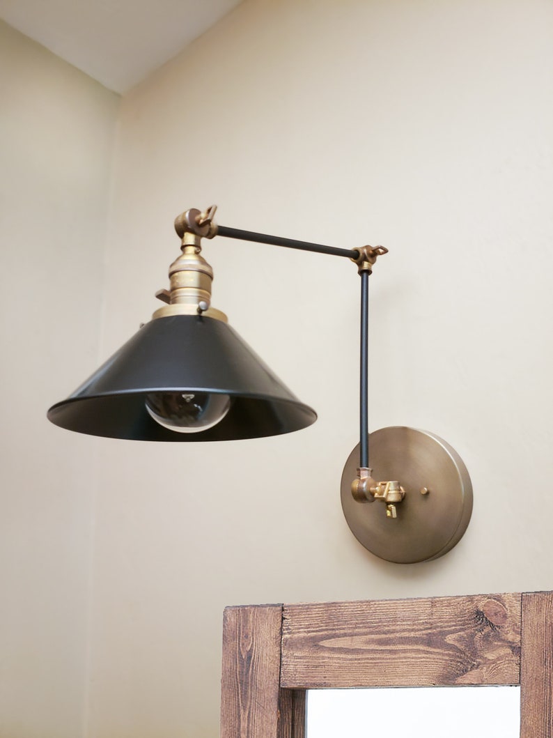 May include: A black and brass wall-mounted light fixture with a black cone-shaped shade and a round brass base. The light is adjustable and can be moved up and down.