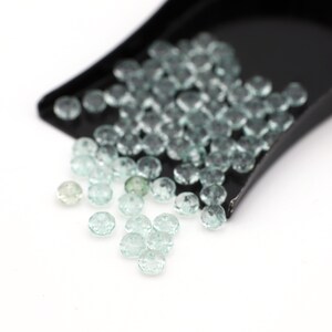 Puede incluir: Una pala llena de piedras preciosas facetadas de color verde claro, con algunas piedras preciosas sueltas esparcidas sobre una superficie blanca.