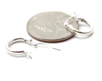 May include: Two small, silver hoop earrings are displayed next to a quarter coin on a white background. The earrings are circular with a hinged closure. The quarter provides a size reference for the earrings.