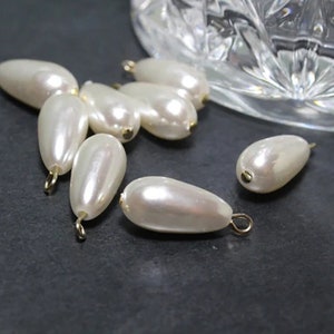 May include: A group of eight white pearlescent teardrop-shaped beads with gold jump rings. The beads are scattered on a dark gray surface.