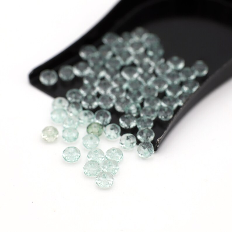 Puede incluir: Una pala llena de piedras preciosas facetadas de color verde claro, con algunas piedras preciosas sueltas esparcidas sobre una superficie blanca.