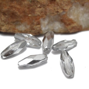 May include: Six clear glass beads with a faceted design. The beads are oval shaped and have a silver finish.