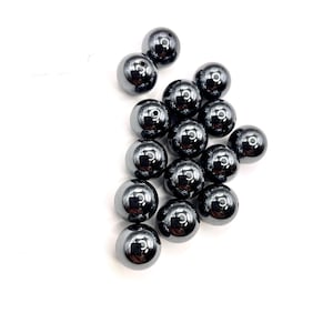 May include: A collection of polished, dark gray hematite beads. Each bead is spherical with a smooth, reflective surface and a small hole for stringing. The beads are scattered against a white background.