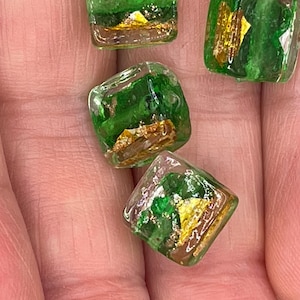 May include: Four square glass beads with green and gold flecks. The beads are translucent and have a slightly iridescent sheen.