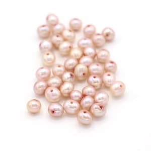 May include: A pile of pink freshwater pearls with a slight iridescent sheen. The pearls are oval shaped and have a small hole drilled through them.