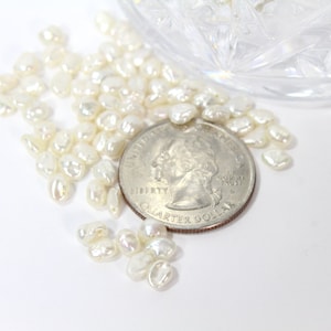 Peut inclure: Un tas de perles d'eau douce de couleur blanche, blanc cassé et crème, certaines avec un éclat iridescent, à côté d'une pièce de 1 euro pour l'échelle.
