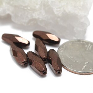 May include: Six faceted, oblong beads in a metallic brown color with a copper-colored accent. A U.S. quarter is included for size comparison. The beads have a faceted surface and are likely for jewelry making.