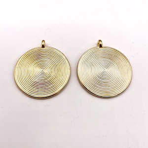 Puede incluir: Dos colgantes circulares dorados con un diseño en espiral. Cada colgante tiene un pequeño lazo en la parte superior para colgar. Los colgantes se muestran sobre un fondo blanco.