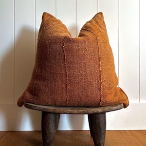 May include: A large, textured, rust-colored pillow with vertical seams, resting on a small, dark wooden stool with three legs. The pillow has a slightly uneven shape and a woven appearance, suggesting a handmade or vintage aesthetic.