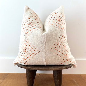May include: A white pillow with an orange geometric pattern sits on a wooden stool. The pillow is made of a textured fabric and has a simple, modern design.