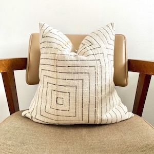 May include: A white throw pillow with a geometric pattern in brown. The pillow is sitting on a brown chair.